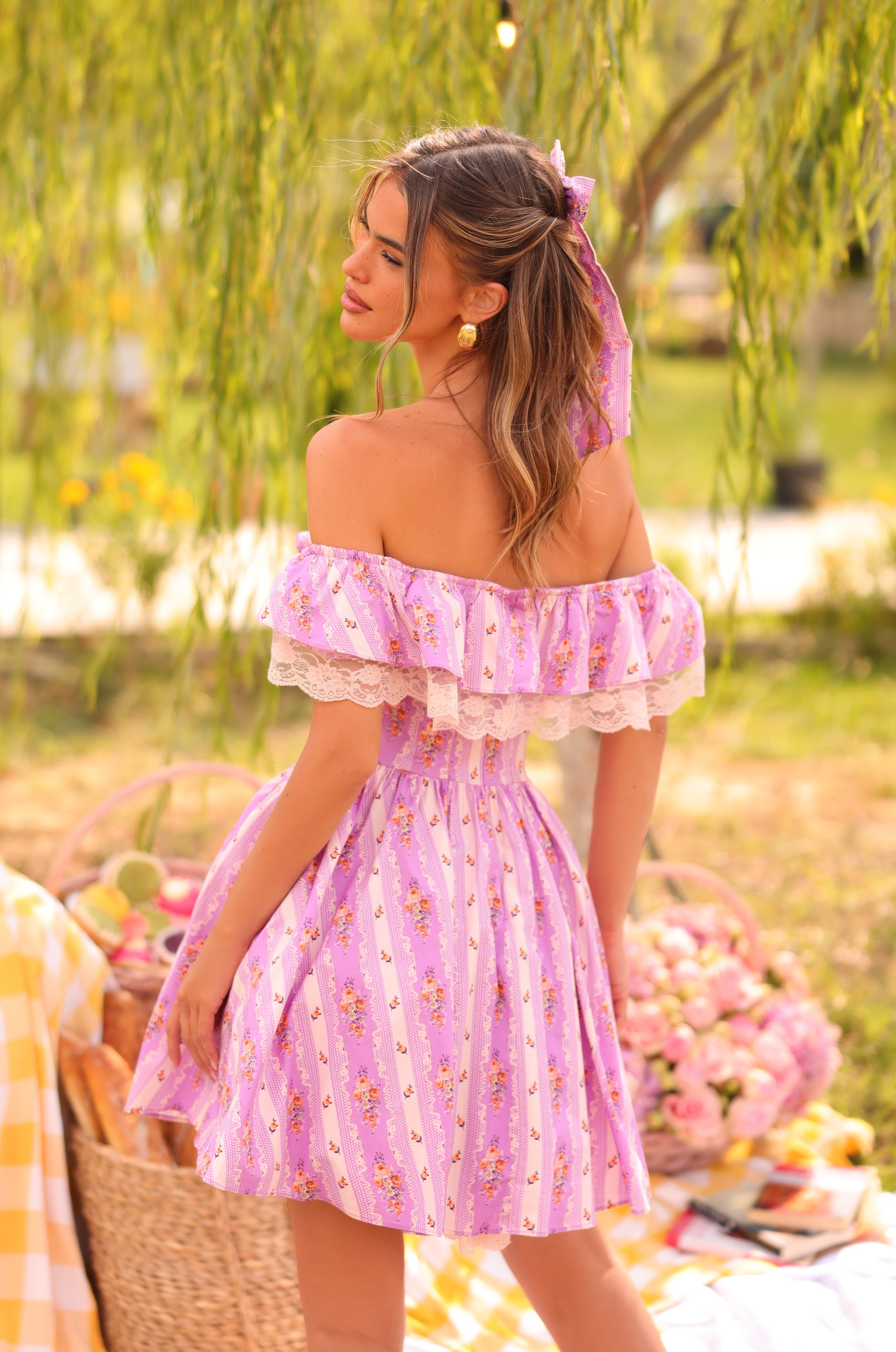 A woman in the Lilac Floral Off-Shoulder Mini Casual Dress: Marella stands outdoors by a picnic setup with baskets, flowers, and a yellow-checkered blanket.